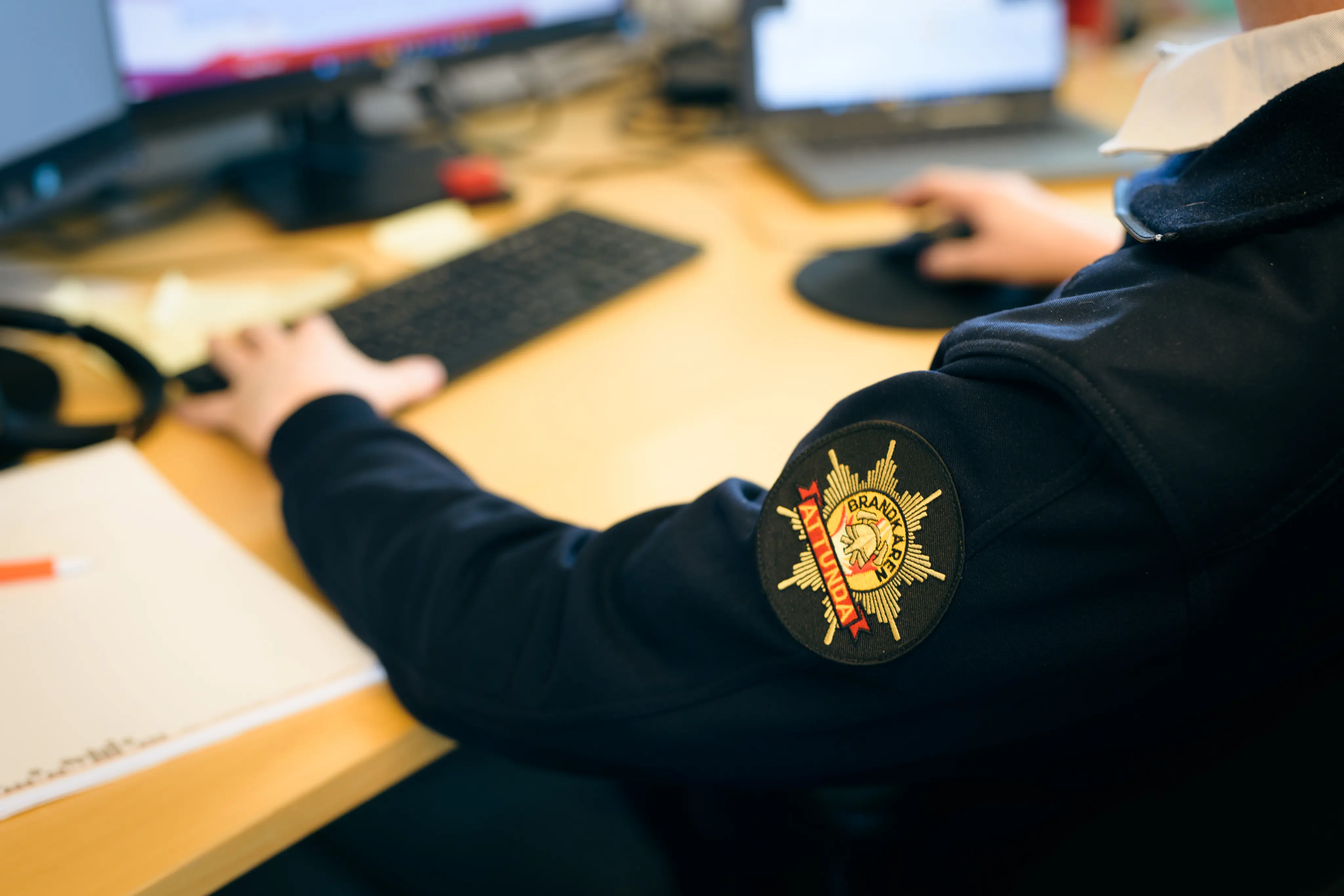 Bild på person vid skrivbord iklädd myndighetströja med Brandkåren Attundas logotyp i förgrunden